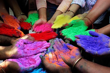 Ten pairs of hands arranged palms-up in a rough circle. Each pair is holding a pile of bright Holi powder